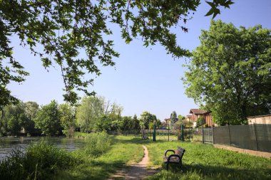 Tahta bank, kısmen gölgede, nehir kıyısında, İtalya kırsalındaki bir parkın yanında.
