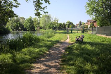 Tahta bank, kısmen gölgede, nehir kıyısında, İtalya kırsalındaki bir parkın yanında.