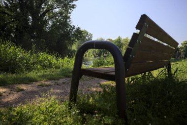 Tahta bank, kısmen gölgede, nehir kıyısında, İtalya kırsalındaki bir parkın yanında.
