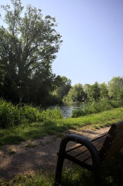 Tahta bank, kısmen gölgede, nehir kıyısında, İtalya kırsalındaki bir parkın yanında.