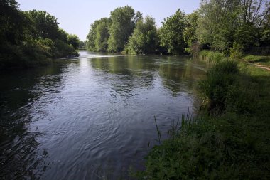 İtalya kırsalındaki bir parktan geçen nehir kıyıdaki ağaçlar ve bitkiler tarafından çerçevelenmiş bir şelale ile.