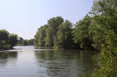 İtalya kırsalındaki bir parktan geçen nehir kıyıdaki ağaçlar ve bitkiler tarafından çerçevelenmiş bir şelale ile.