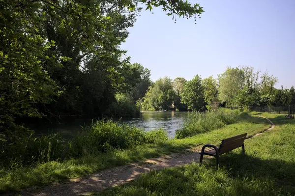 Tahta bank, kısmen gölgede, nehir kıyısında, İtalya kırsalındaki bir parkın yanında.