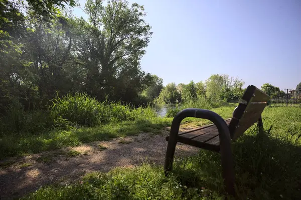 Tahta bank, kısmen gölgede, nehir kıyısında, İtalya kırsalındaki bir parkın yanında.
