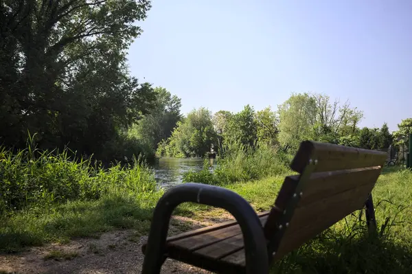 Tahta bank, kısmen gölgede, nehir kıyısında, İtalya kırsalındaki bir parkın yanında.