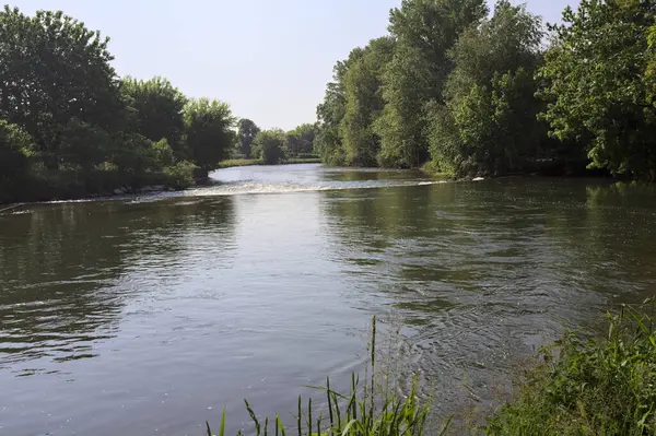 İtalya kırsalındaki bir parktan geçen nehir kıyıdaki ağaçlar ve bitkiler tarafından çerçevelenmiş bir şelale ile.