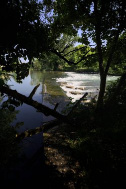 İtalya kırsalındaki bir parkta, üzerinde şelale olan bir nehirde kıvrıl. Gölgedeki ağaçlarla çerçevelenmiş.