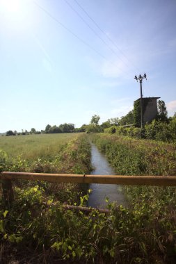 İtalya kırsalında terk edilmiş bir yerleşkenin kenarındaki çukur üzerinde gelincikler yetişen paslı bir trabzanın arkasından görülüyor.