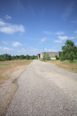 Terk edilmiş bir depo ve önünde çıplak bir avludan görünen asfaltlı bir giriş yolu var.