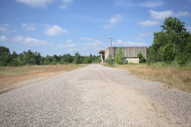 Terk edilmiş bir depo ve önünde çıplak bir avludan görünen asfaltlı bir giriş yolu var.