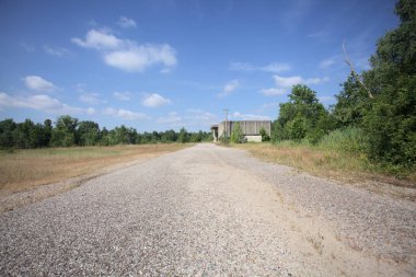 Terk edilmiş bir depo ve önünde çıplak bir avludan görünen asfaltlı bir giriş yolu var.