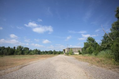 Terk edilmiş bir depo ve önünde çıplak bir avludan görünen asfaltlı bir giriş yolu var.