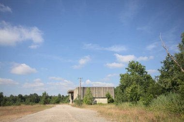 Terk edilmiş bir depo ve önünde çıplak bir avludan görünen asfaltlı bir giriş yolu var.