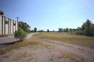 Terk edilmiş bir depo ve önünde çıplak bir avludan görünen asfaltlı bir giriş yolu var.