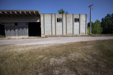 Terk edilmiş bir yerleşkenin depoları güneşli bir günde ağaçlarla çevrili İtalya kırsalında kuru çimenler ve çakıllarla çevrili bir bahçeden görülebiliyor.
