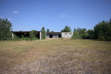 Terk edilmiş bir yerleşkenin depoları güneşli bir günde ağaçlarla çevrili İtalya kırsalında kuru çimenler ve çakıllarla çevrili bir bahçeden görülebiliyor.