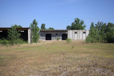 Terk edilmiş bir yerleşkenin depoları güneşli bir günde ağaçlarla çevrili İtalya kırsalında kuru çimenler ve çakıllarla çevrili bir bahçeden görülebiliyor.