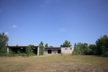 Terk edilmiş bir yerleşkenin depoları güneşli bir günde ağaçlarla çevrili İtalya kırsalında kuru çimenler ve çakıllarla çevrili bir bahçeden görülebiliyor.