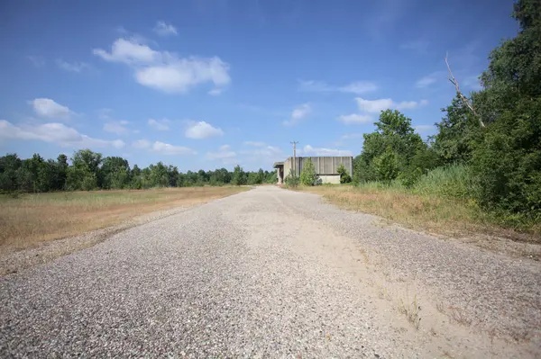 Terk edilmiş bir depo ve önünde çıplak bir avludan görünen asfaltlı bir giriş yolu var.