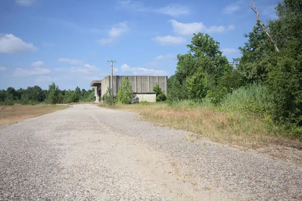 Terk edilmiş bir depo ve önünde çıplak bir avludan görünen asfaltlı bir giriş yolu var.
