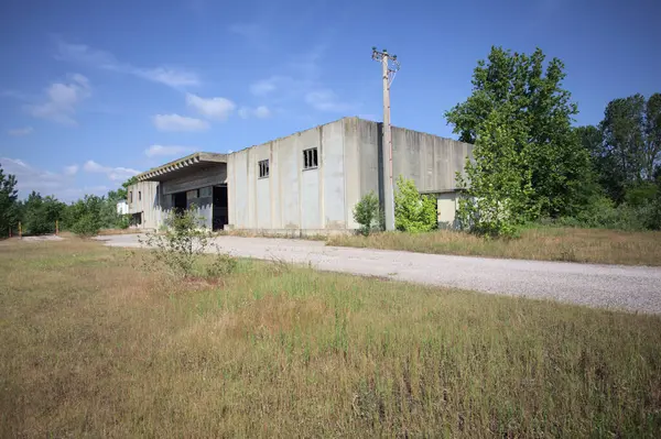 Terk edilmiş bir depo ve önünde çıplak bir avludan görünen asfaltlı bir giriş yolu var.
