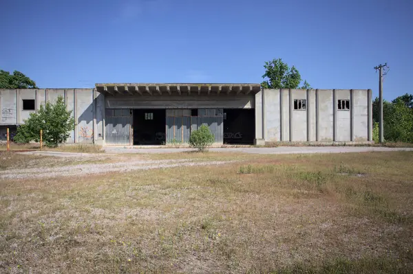 Terk edilmiş bir yerleşkenin depoları güneşli bir günde ağaçlarla çevrili İtalya kırsalında kuru çimenler ve çakıllarla çevrili bir bahçeden görülebiliyor.