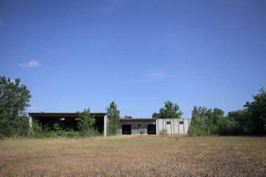 Terk edilmiş bir yerleşkenin depoları güneşli bir günde ağaçlarla çevrili İtalya kırsalında kuru çimenler ve çakıllarla çevrili bir bahçeden görülebiliyor.