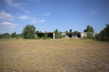 Terk edilmiş bir yerleşkenin depoları güneşli bir günde ağaçlarla çevrili İtalya kırsalında kuru çimenler ve çakıllarla çevrili bir bahçeden görülebiliyor.