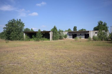 Terk edilmiş bir yerleşkenin depoları güneşli bir günde ağaçlarla çevrili İtalya kırsalında kuru çimenler ve çakıllarla çevrili bir bahçeden görülebiliyor.