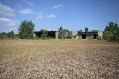 Terk edilmiş bir yerleşkenin depoları güneşli bir günde ağaçlarla çevrili İtalya kırsalında kuru çimenler ve çakıllarla çevrili bir bahçeden görülebiliyor.