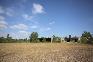 Terk edilmiş bir yerleşkenin depoları güneşli bir günde ağaçlarla çevrili İtalya kırsalında kuru çimenler ve çakıllarla çevrili bir bahçeden görülebiliyor.