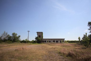 Çalıların yanındaki terk edilmiş bina ve güneşli bir günde çıplak bir avludan görülen elektrik hattı.