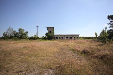 Çalıların yanındaki terk edilmiş bina ve güneşli bir günde çıplak bir avludan görülen elektrik hattı.