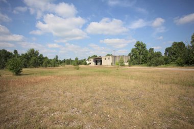 Terk edilmiş bir yerleşkenin depoları güneşli bir günde ağaçlarla çevrili İtalya kırsalında kuru çimenler ve çakıllarla çevrili bir bahçeden görülebiliyor.