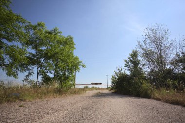 Eskimiş asfalt yollar, güneşli bir günde ağaçların çerçevelediği paslı bir bar tarafından kapatılmış.