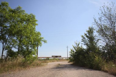 Eskimiş asfalt yollar, güneşli bir günde ağaçların çerçevelediği paslı bir bar tarafından kapatılmış.