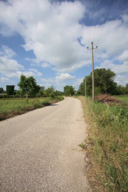 İtalya 'nın kırsal kesimlerinde güneşli bir günde bir bar tarafından engellenmiş, üzerinde elektrik hattı olan tarlalar arasındaki kirli yol.