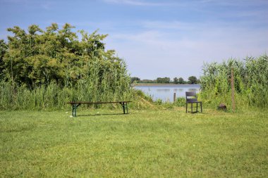 Güneşli bir günde göl kenarındaki bankta ve sandalyede sazlıkların çerçevelediği karşı kıyıda.
