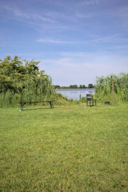 Güneşli bir günde göl kenarındaki bankta ve sandalyede sazlıkların çerçevelediği karşı kıyıda.