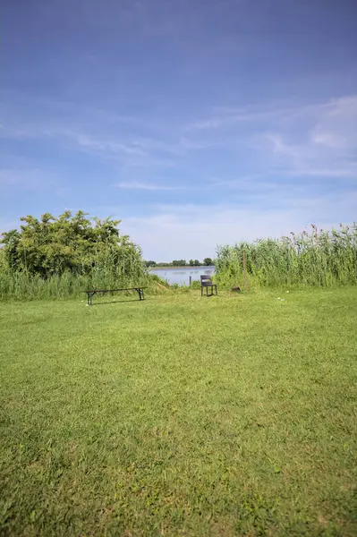 Güneşli bir günde göl kenarındaki bankta ve sandalyede sazlıkların çerçevelediği karşı kıyıda.