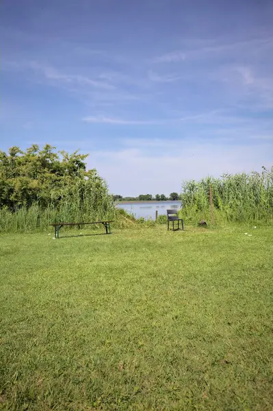 Güneşli bir günde göl kenarındaki bankta ve sandalyede sazlıkların çerçevelediği karşı kıyıda.