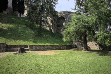 Etrafını çevreleyen ağaçlarla çevrili çimenler ve ağaçların arkasında bir şatonun kalıntıları olan gölgeler.