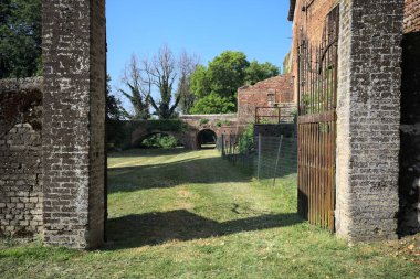 Bir şatonun kenarındaki patika, uzaktaki bir köprünün kemerlerinin altından geçiyor, yere düşen ağaçların gölgeleriyle.