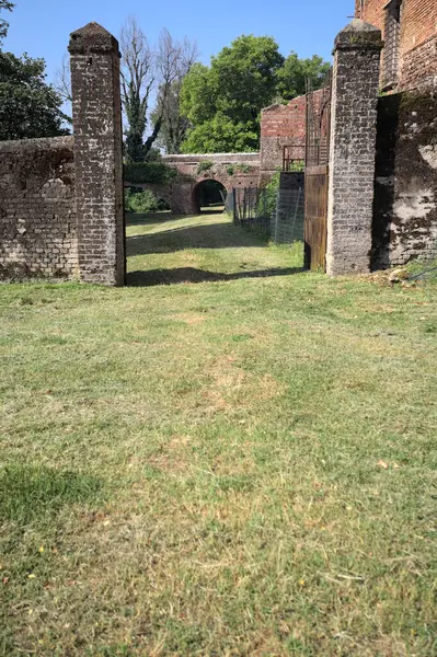 Bir şatonun kenarındaki patika, uzaktaki bir köprünün kemerlerinin altından geçiyor, yere düşen ağaçların gölgeleriyle.