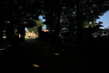 Güneşli bir günde, İtalya kırsalında bir parka açılan açık kapılı giriş kapısı. Kapının yapısı çerçeveli ve gölgede ağaçlar var.