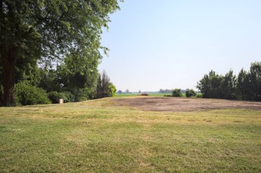 İtalya 'nın kırsal kesimlerinde güneşli bir günde bitkilerden yapılmış bir çitin arkasında ufka kadar uzanan bir mısır tarlası olan ağaçlarla çevrili beton kare.