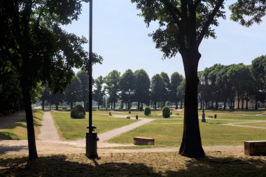 Bir köşeden görünen ve ağaçların ve bankların siluetleriyle çerçevelenmiş kavak ağaçlarıyla çevrili bir meydan.