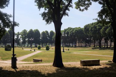 Bir köşeden görünen ve ağaçların ve bankların siluetleriyle çerçevelenmiş kavak ağaçlarıyla çevrili bir meydan.