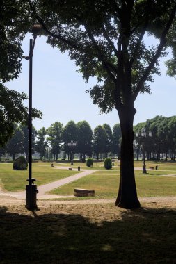Bir köşeden görünen ve ağaçların ve bankların siluetleriyle çerçevelenmiş kavak ağaçlarıyla çevrili bir meydan.
