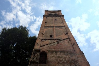 Arka plan olarak kısmen bulutlu bir gökyüzü olan ağaçların yanındaki tuğla çan kulesinin kalıntıları.